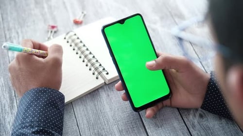 Rückansicht eines jungen Mannes, der Smartphone benutzt und auf Notizblock schreibt