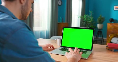 Man Uses Laptop with Green Mockup Screen While Sitting at the Desk in His Cozy