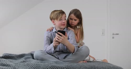 Boy Looking at Phone with Sister on Bed