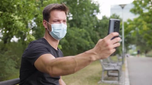 A Young Man Takes Selfies with a Smartphone As He Sits on a Bench in a Park - Closeup From the Side
