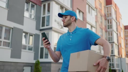 A Male Mail Carrier with Round Glasses Carries Boxes for Delivery to Customers and Looks at His
