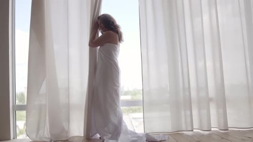 Attractive Woman Covered in the Bedsheet Standing Near the Floor-to-ceiling Window and Looking Away