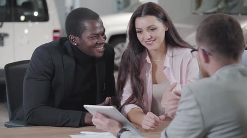 Vista traseira do jovem concessionário de carros caucasiano mostrando automóveis em seu tablet para jovem multirracial