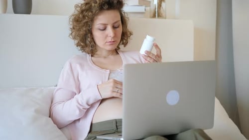Pregnant Woman Holds Pills While Using Laptop