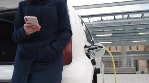 Business Woman Standing Near Charging Electric Car or EV Car and Using Mobile Phone in the Street