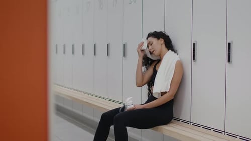 Young Tired Sporty Lady Resting at Gym Locker Room Wiping Her Forehead with Towel Sitting on Bench