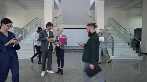 Caucasian Man and Woman Discussing Business Plan Standing in Office Lobby with Employees Passing