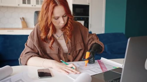 Rothaarige attraktive Mädchen-Designerin mit einer Prothese arbeitet an einem Projekt in ihrem Heimbüro