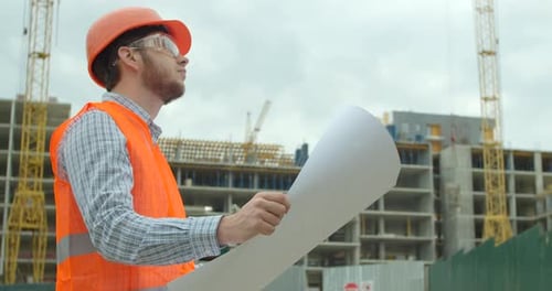 Young Architect or Builder With Hard Hat Holding Blueprint In His Hands in Front of Building Under
