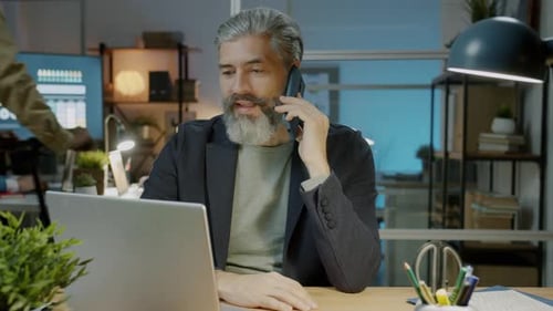 Mature Office Worker Speaking on Mobile Phone Working at Night Concentrated on Business Conversation
