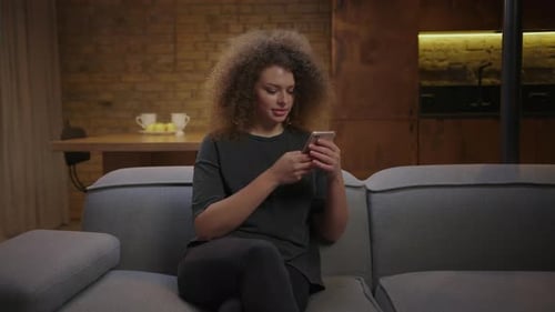 Young Adult Woman Using Cell Phone for Texting with Friends Sitting on Couch at Home