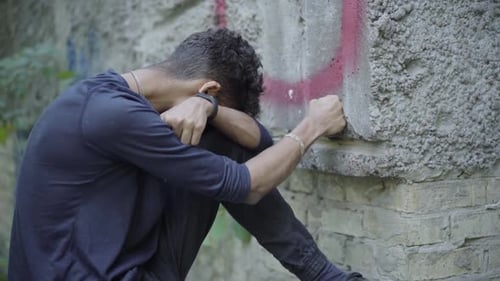 Verzweifelter einsamer junger Mann, der mit der Faust die Wand schlägt und weint. Seitenansicht von frustriert gestresst