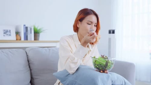 Asian attractive woman sit on sofa, eating green salad in living room.