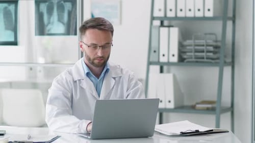 Positive Male Doctor Working on Laptop at Desk and Posing for Camera