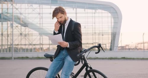 Hombre que está de pie cerca de su bicicleta en el edificio de oficinas y tiene una conversación telefónica