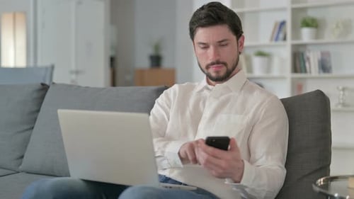 Man Using Laptop and Smartphone on Couch Indoors