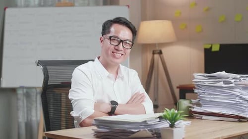 Asian Man Crossing Arms And Smiling To Camera While Working With Documents At The Office