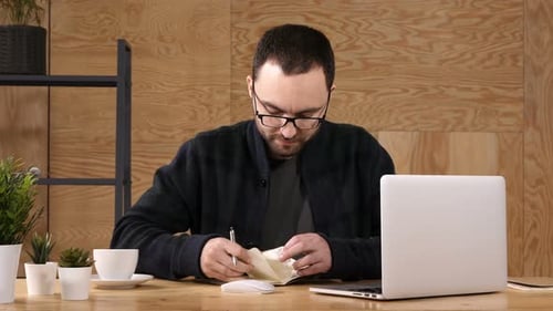 Man Writing in Notebook at Desk with Laptop