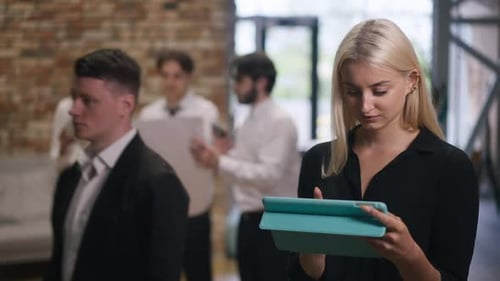 Smiling Beautiful Woman Analyzing Project on Tablet Standing in Office Hall with Colleagues Passing