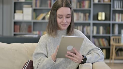 Woman on Couch Using Tablet in Front of Bookshelf