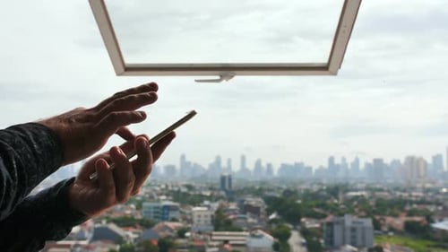 A Man Types a Message on a Smartphone Against the Background of an Open Window and a Panorama of the