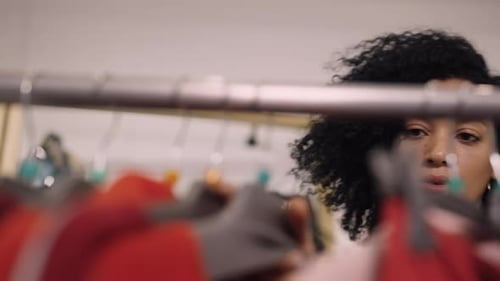 Beautiful African American Woman Choosing Clothes Hanging on Rack Hangers in a Trendy Clothing Store