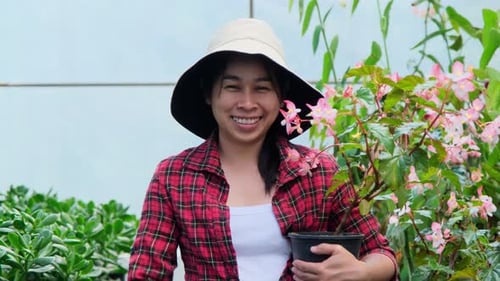 Gardener woman holding small plant pot and looking at camera in greenhouse.