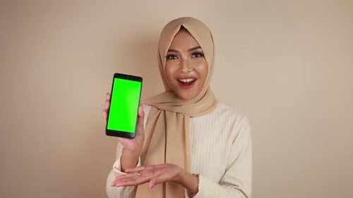 Happy young Muslim Asian woman pointing to a cellphone with green screen