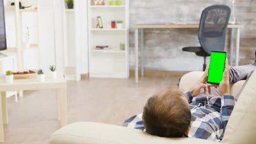 Parallax Shot of Male Lying on the Sofa and Looking at Green Screen Phone