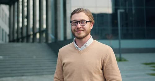 Empresario en gafas sonriendo a la cámara mientras está de pie al aire libre junto al edificio de oficinas