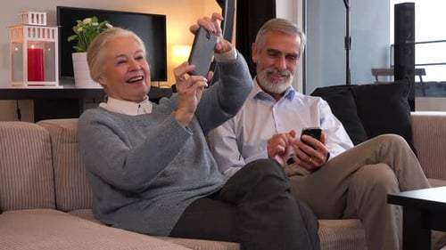 An Elderly Couple Sits on A Couch in An Apartment and Each Works on And Plays with Their Smartphone