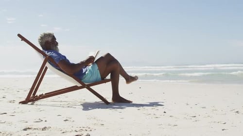 Senior african american man reading and lying on sunbed on sunny beach