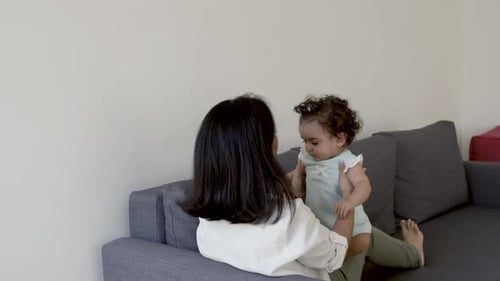 Back View of Mom Lying on Sofa Lifting Cute Daughter Over Head