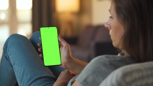 Woman at Home Lying on a Sofa and Using Smartphone with Green Mock-up Screen in Vertical Mode. Girl