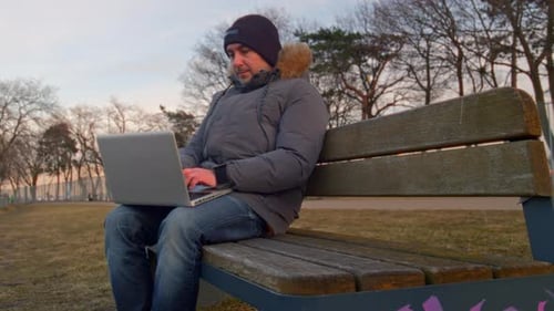 Man is Working on a Laptop While Sitting on a Park Bench