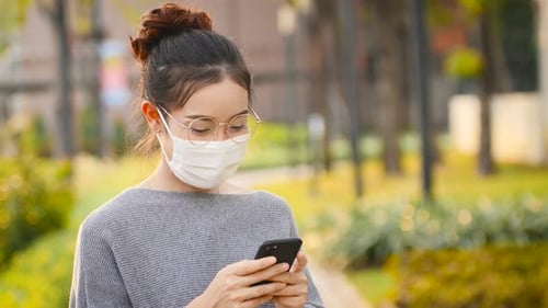 Woman Wearing Face Mask Uses Cellphone Outdoors