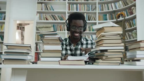 Glücklicher afrikanischer Student, der Musik hört, während er am Tisch in der Bibliothek sitzt