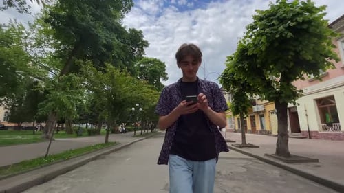 Calm Millenial Hipster Guy Holding Smartphone in His Hand Walking Down the Street Old City
