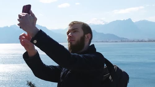 A young bearded male tourist takes a selfie on a mobile phone against the backdrop of a bay