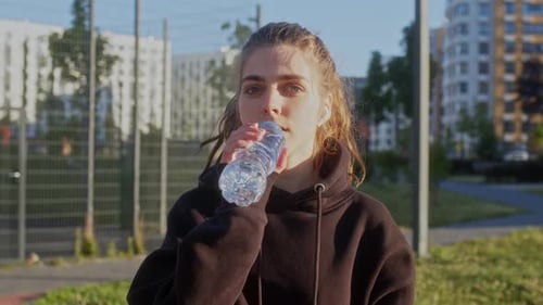 Young Woman Sits on Bench in Apartment Complex in Morning After Jogging and Drinks Water