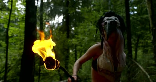 Masked Woman Holds Flaming Torch in Green Forest
