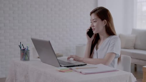 Mulher asiática de negócios Fale com o trabalho ao telefone e faça anotações no laptop com o trabalho em casa.
