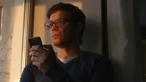 Cute Man Looks To The Mobile Phone And Then To The Window At The Sunset