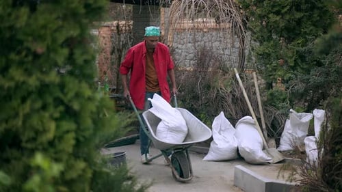 Wide Shot of African American Man Pushing Construction Trolley with Broken Wheel Outdoors in Garden