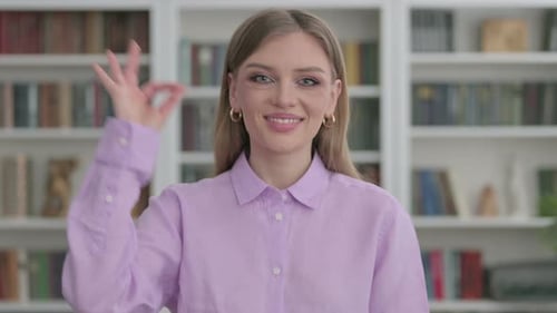 Young Woman Makes OK Sign in Front of Bookshelf