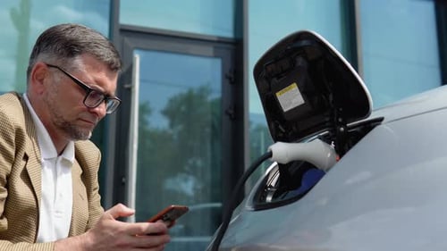 Man charging electric car with phone outdoors