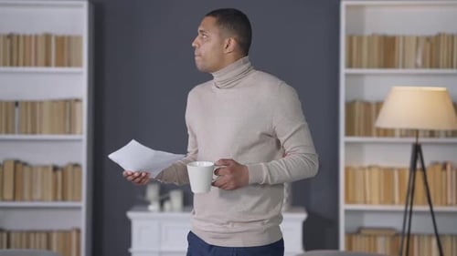 Unruhiger afroamerikanischer junger Mann steht im Wohnzimmer mit Kaffeetasse und Dokumenten
