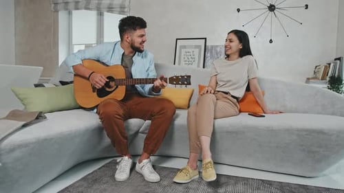 Young Man Plays Guitar for Smiling Friend on Couch