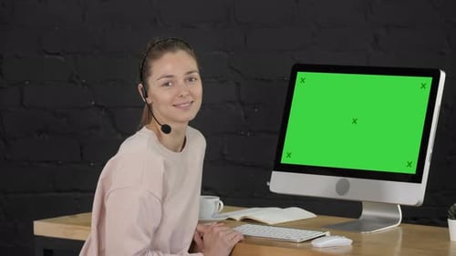 Lächelnde Frau blickt mit Headset und Computermonitor in die Kamera. Greenscreen-Mockup-Display.