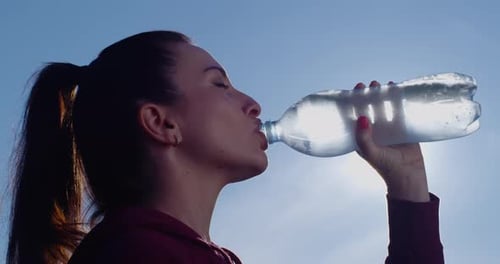 Perfil de mujer hermosa bebiendo agua de una botella afuera con sol retroiluminado y cielo azul